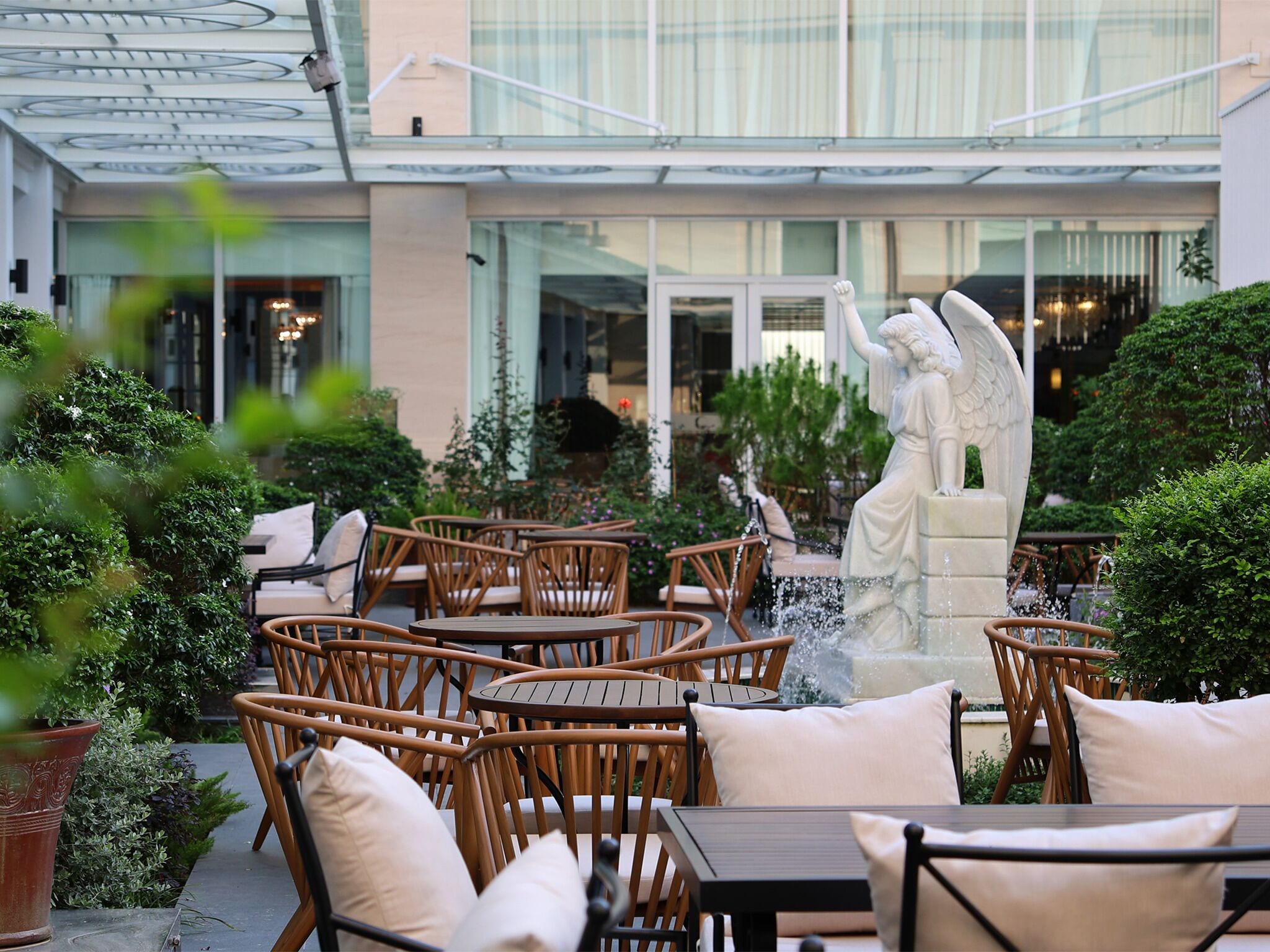 Een terras buiten met rieten stoelen en tafels, een standbeeld van een engel en een fontein.