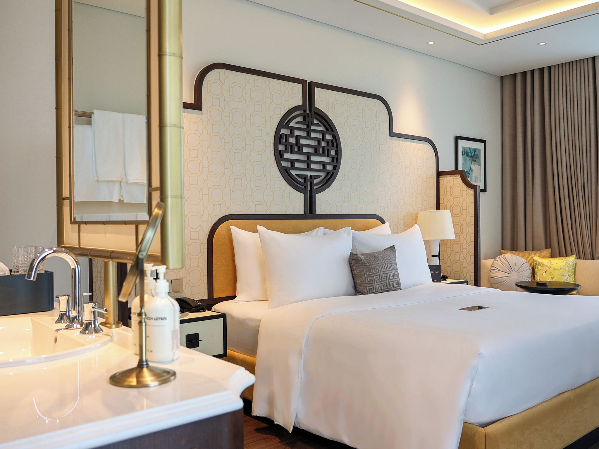A hotel room with a large bed, a bedside table with a lamp, a nightstand, a bathroom with a sink and mirror, and a decorative wall panel.