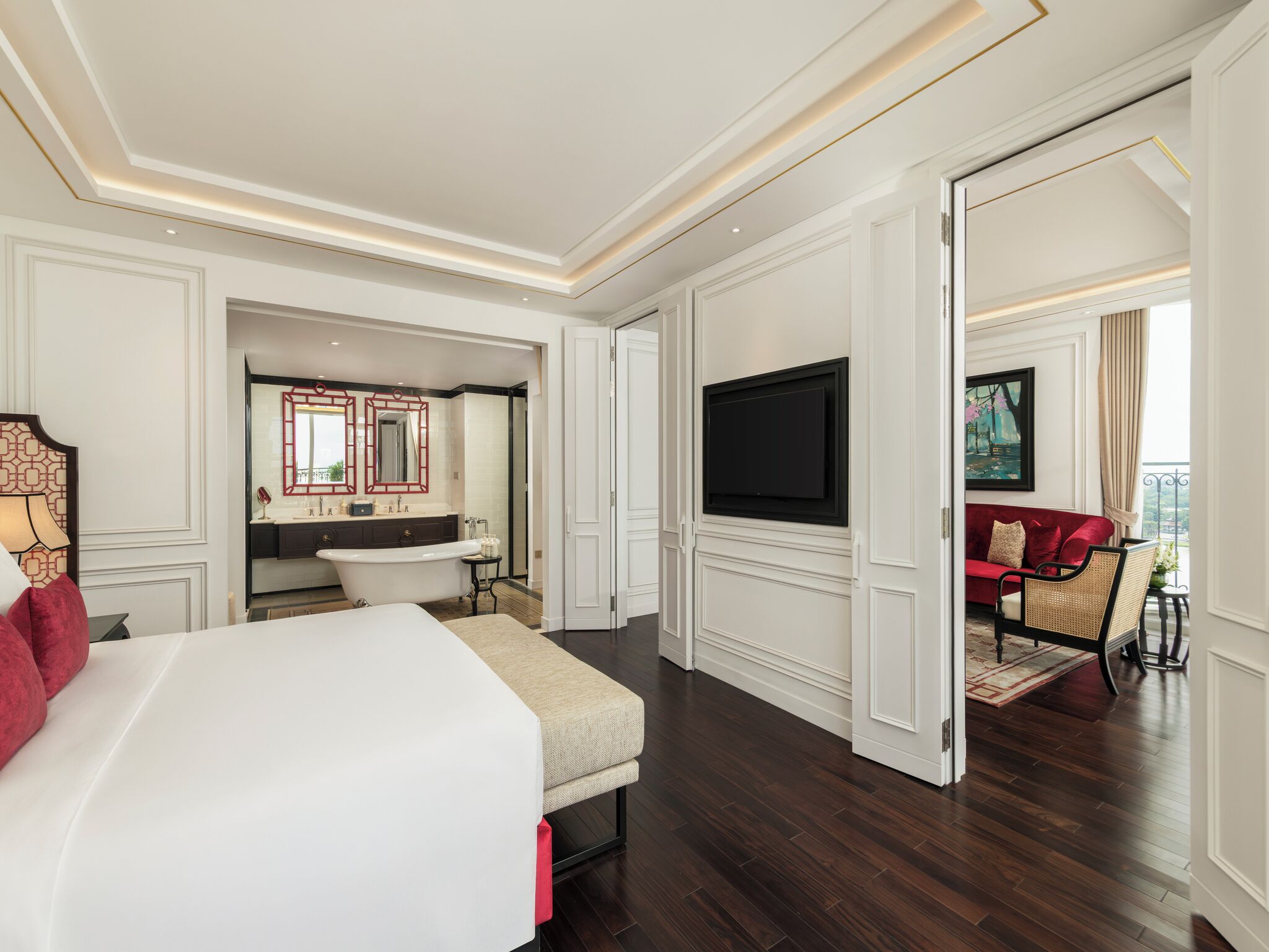 A hotel room with a large bed, a sitting area with a red sofa, a television mounted on the wall, and a bathroom with a bathtub and mirrors.