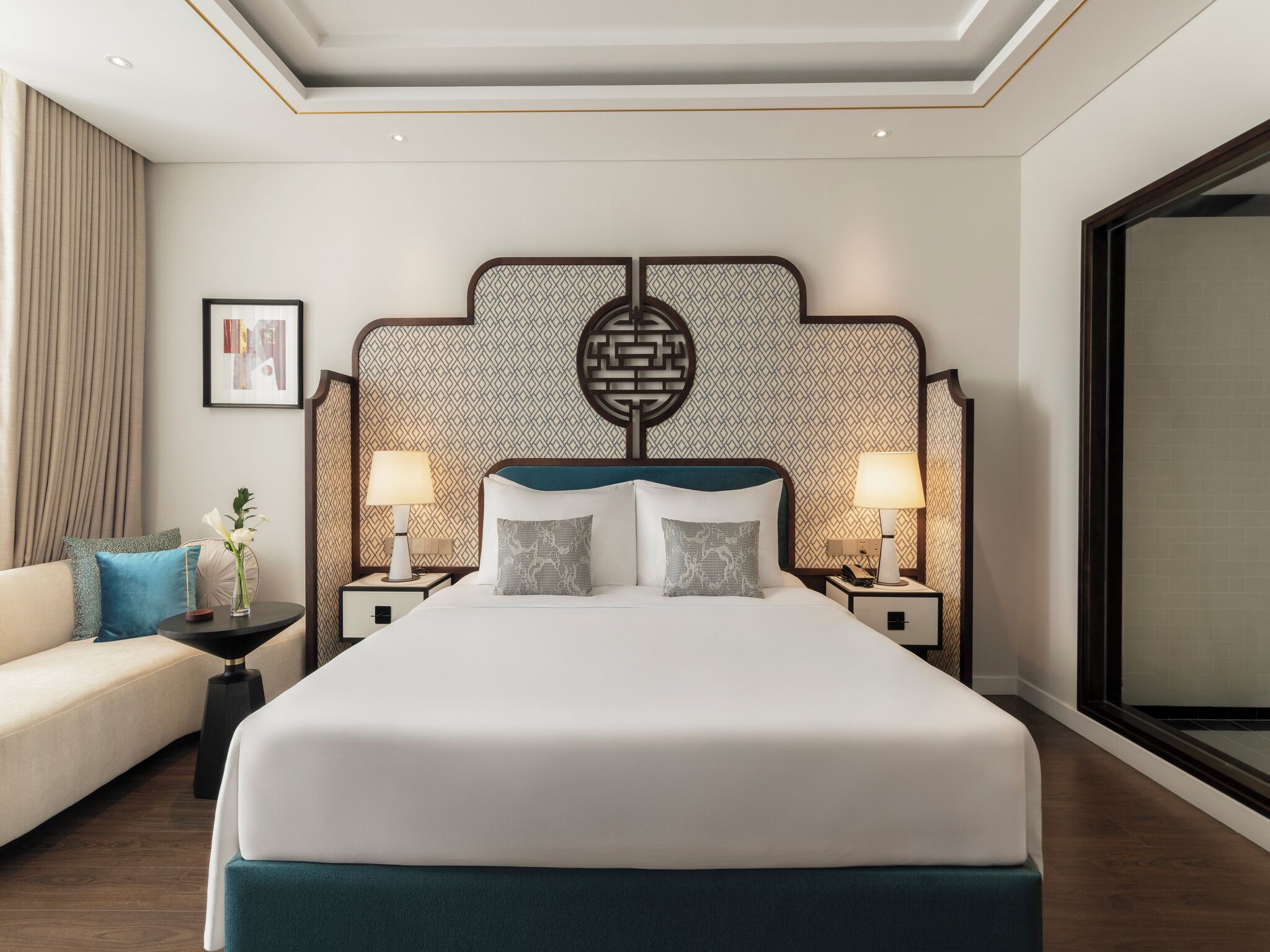 A hotel room with a large bed, two bedside tables with lamps, a nightstand, a sofa, and a large window.