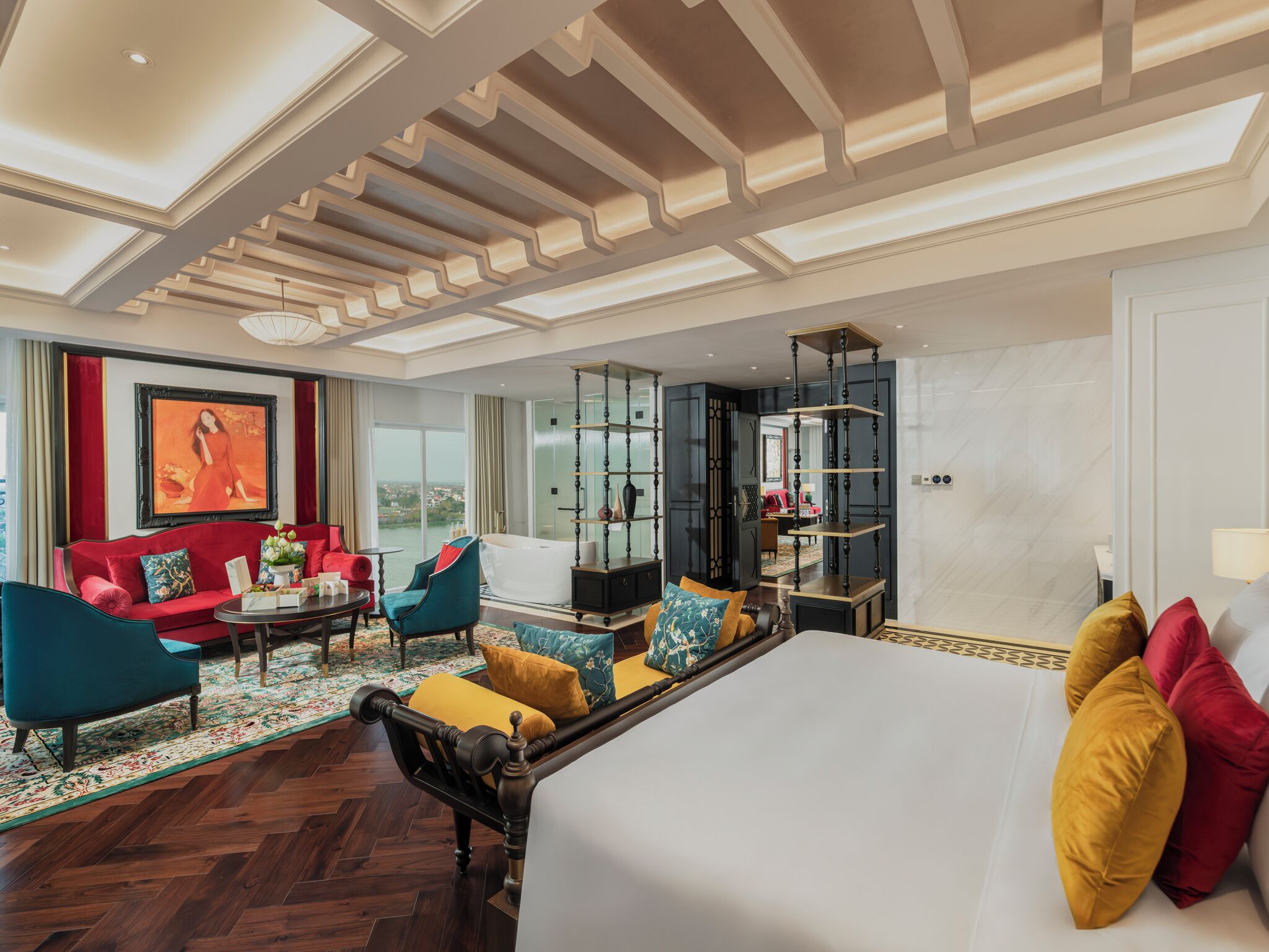 A hotel room with a large bed, a sitting area with a sofa and chairs, a dining table, a large window with a view, and a decorative ceiling.