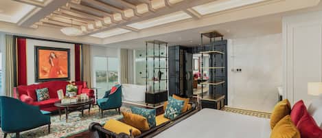 A hotel room with a large bed, a sitting area with a sofa and chairs, a dining table, a large window with a view, and a decorative ceiling.