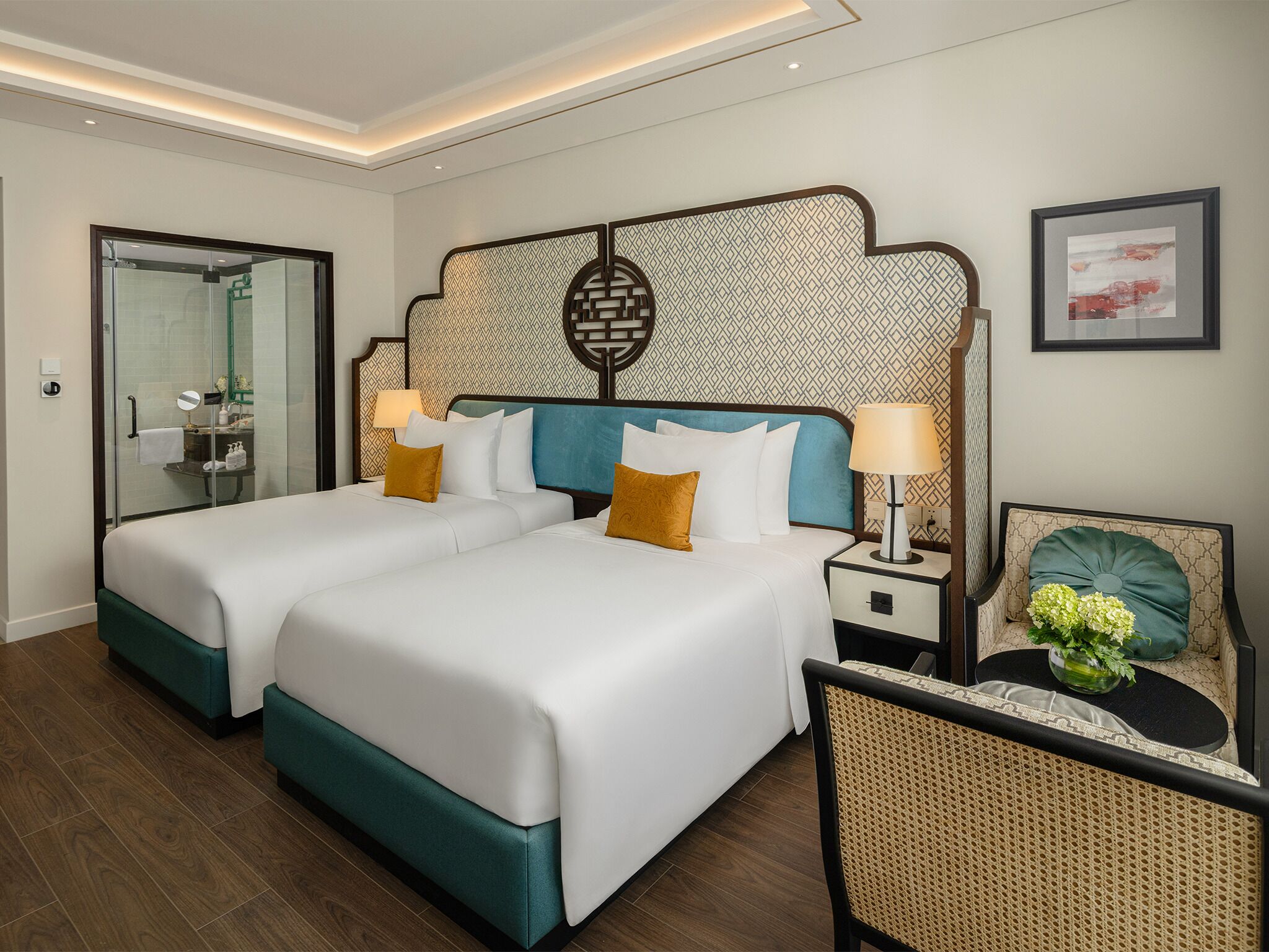 A hotel room with two beds, a bathroom with a glass door, a bedside table with a lamp, and a chair with a vase of flowers.