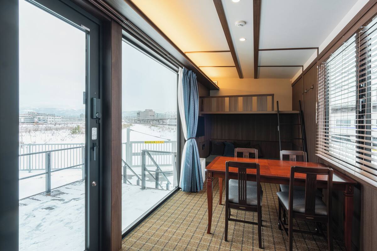 A room with a large sliding glass door leading to a snowy outdoor area, a wooden dining table with chairs, and a small kitchen area with wooden cabinets and a window with blinds.