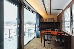 A room with a large sliding glass door leading to a snowy outdoor area, a wooden dining table with chairs, and a small kitchen area with wooden cabinets and a window with blinds. - JR Mobile Inn Hakodate (Hakodate)