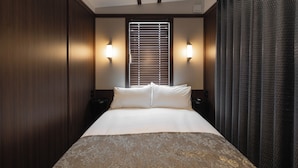 Une chambre d’hôtel avec un grand lit, deux tables de chevet, une fenêtre avec des stores et un rideau foncé.