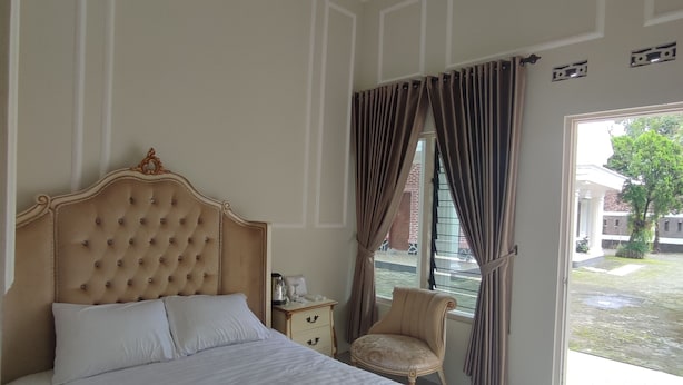 Una cama grande con cabecera capitonada, una mesita de noche con lámpara y una silla junto a una ventana con cortinas.