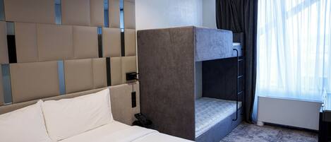 A hotel room with a large bed, a bunk bed, a bedside table, a telephone, and a window with curtains.