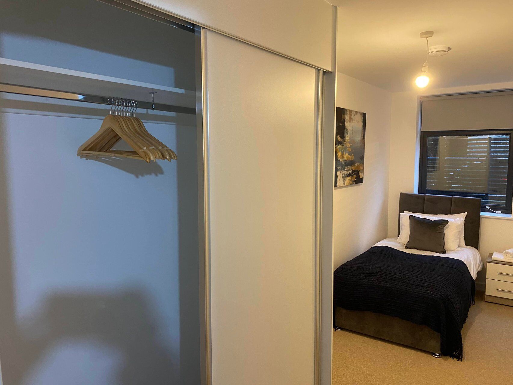 A bedroom with a bed, a nightstand, a window with blinds, a painting on the wall, and a closet with a hanging rack.
