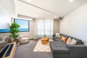 Uma sala de estar moderna com um sofá cinzento, uma poltrona branca, uma mesa de centro de madeira e uma janela ampla com vista para o mar.
