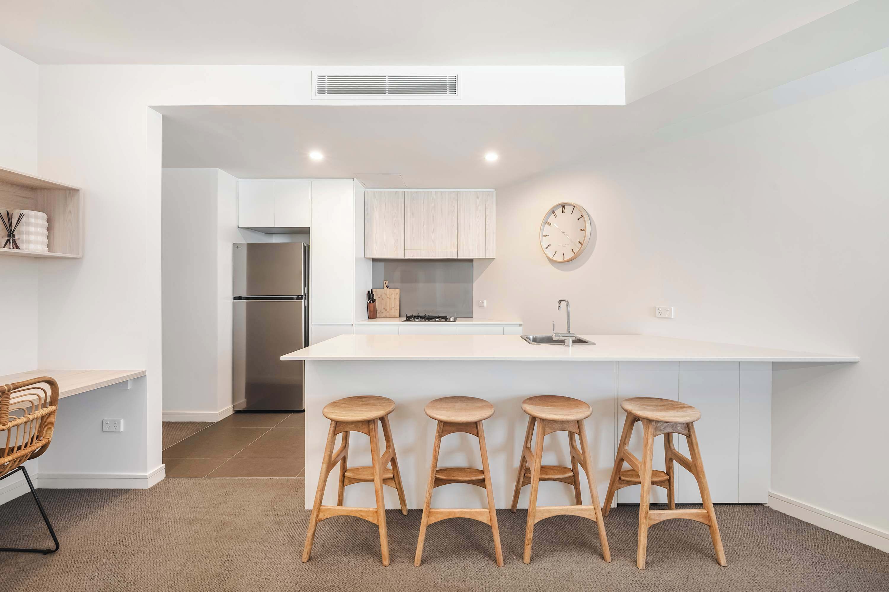 A modern kitchen with a white island, three wooden stools, a stainless steel refrigerator, and a clock on the wall.