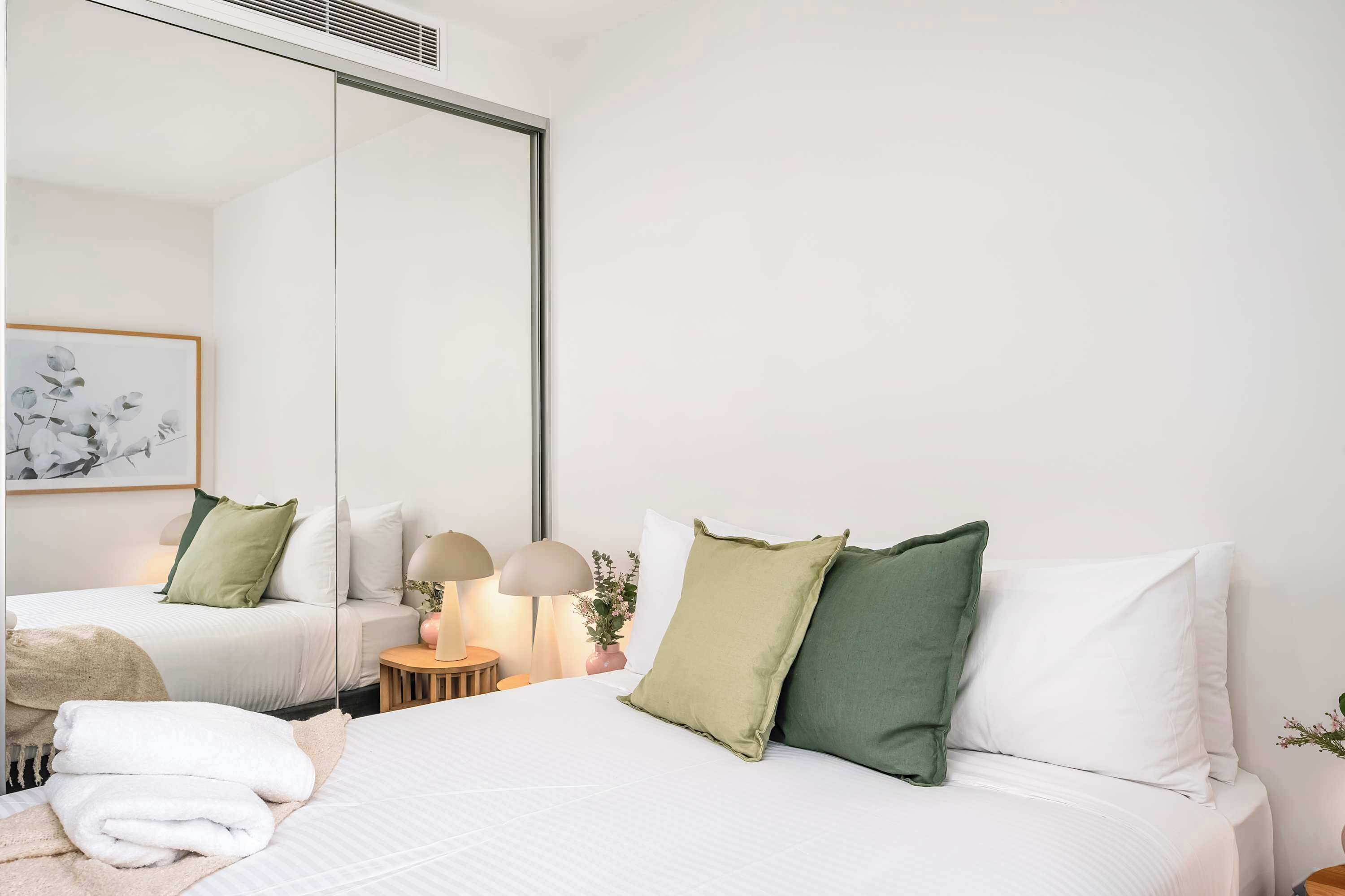 A hotel room with two beds, a large mirror, a side table with lamps, and a framed artwork on the wall.