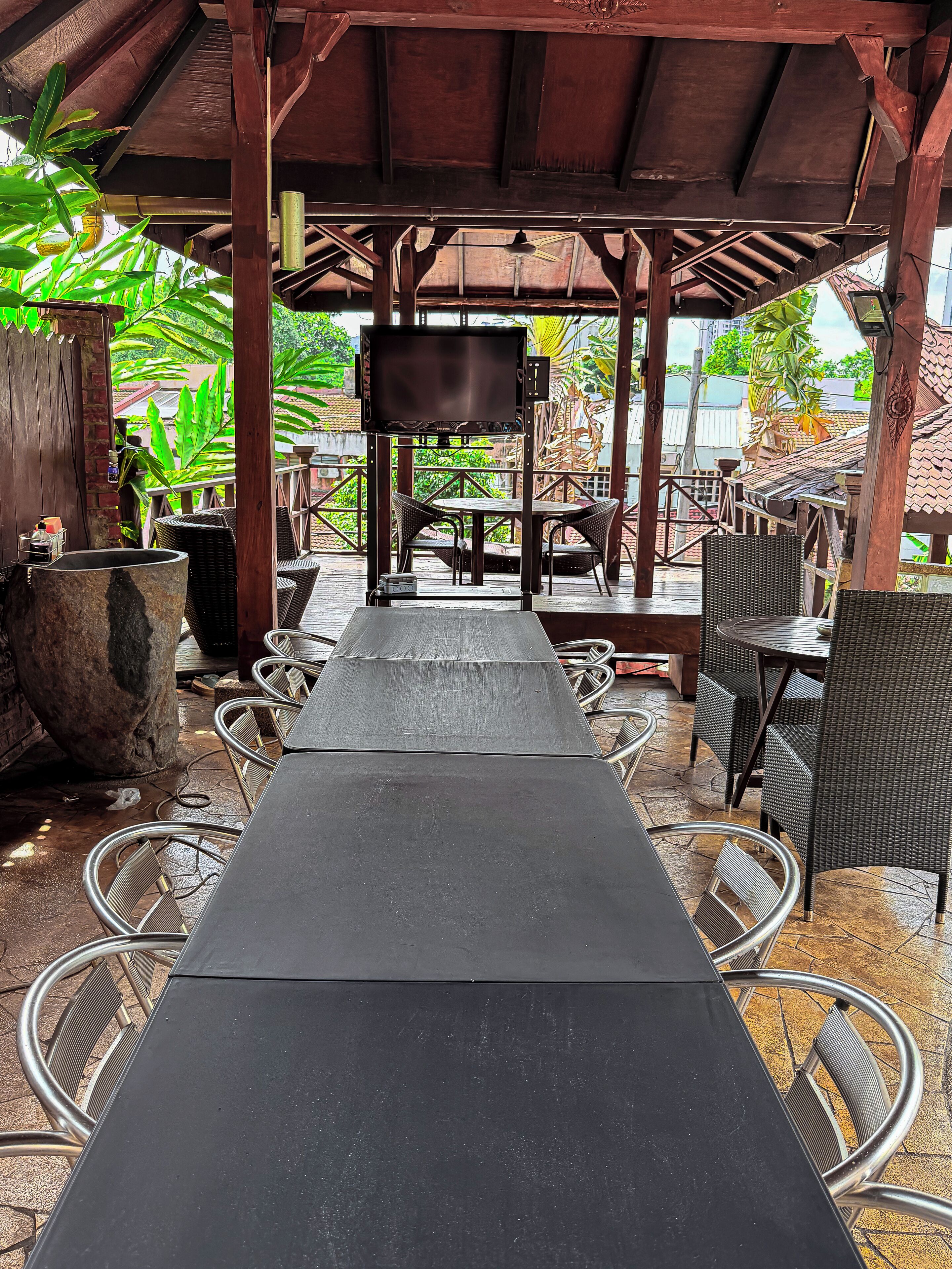 A covered outdoor seating area with a long table and chairs, a television screen, and a stone planter.