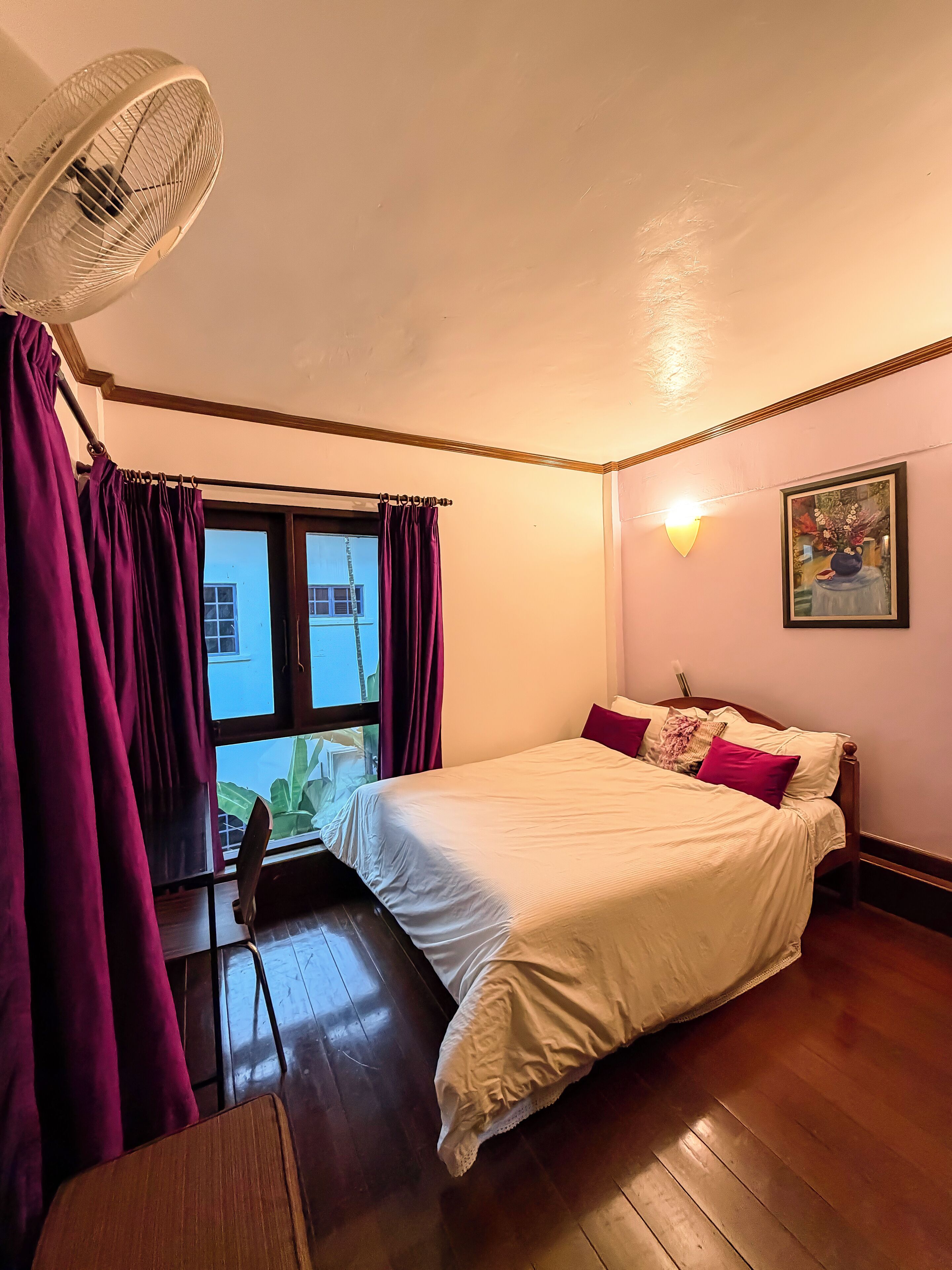 A bedroom with a bed, a window with curtains, a fan, and a painting on the wall.