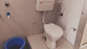 Ένα μικρό μπάνιο με μια τουαλέτα, ένα μπλε κουβά και μια λευκή δεξαμενή νερού.