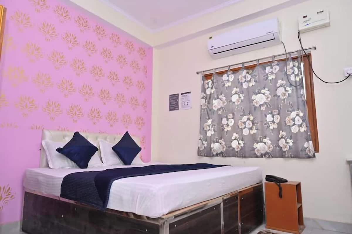 A bedroom with a bed, a window with floral curtains, an air conditioning unit, and a wall-mounted air conditioner.