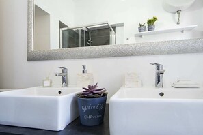 Un baño con dos lavamanos blancos, un espejo grande y una planta en maceta, con la etiqueta 'Garden Life Collection', ubicada entre ambos.