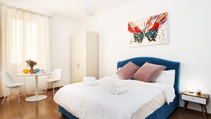 Moderni hotellihuone, jossa on suuri sänky, yöpöytä lampun kanssa, vaatekaappi, pyöreä ruokapöytä tuoleineen sekä värikäs abstrakti maalaus seinällä.