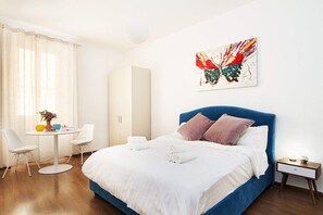 Ein modernes Hotelzimmer mit einem großen Bett, einem Nachttisch mit Lampe, einem Kleiderschrank, einem runden Esstisch mit Stühlen und einem farbenfrohen, abstrakten Gemälde an der Wand.