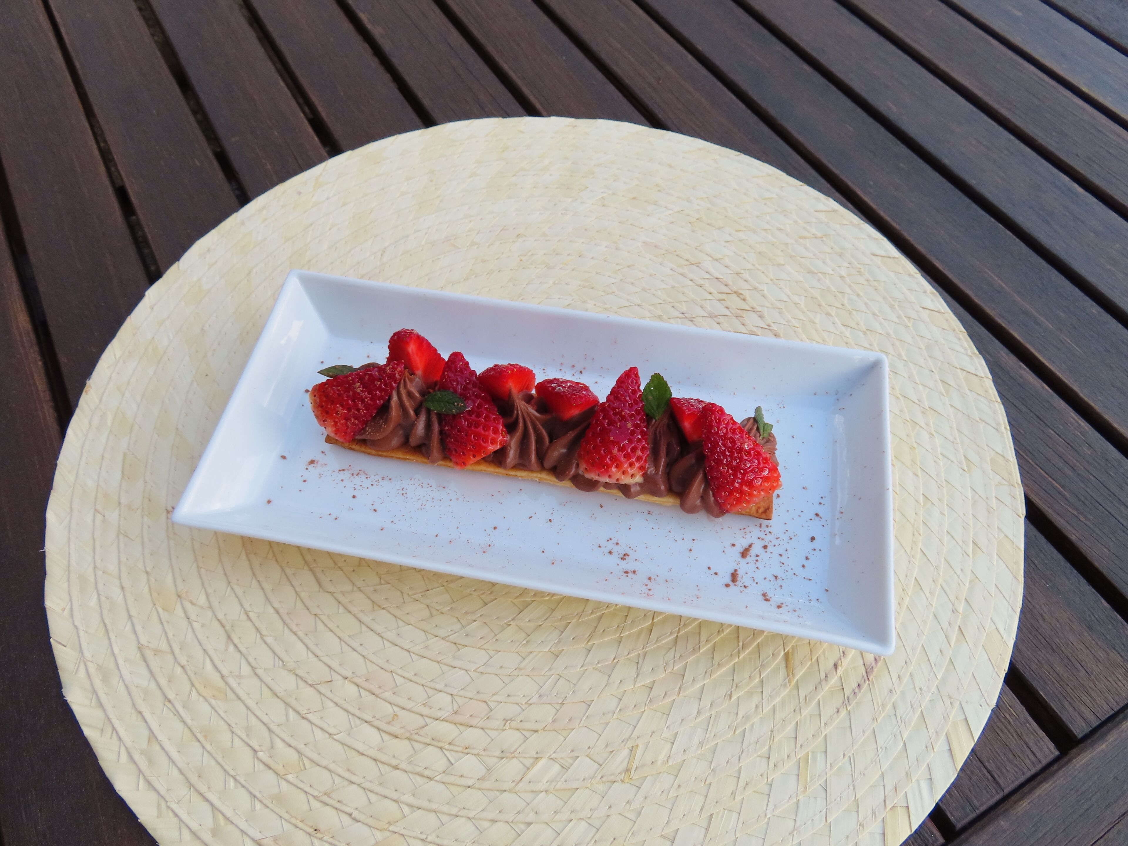 Un plato rectangular blanco con un postre de chocolate cubierto de fresas y hojas de menta, sobre un tapete tejido.