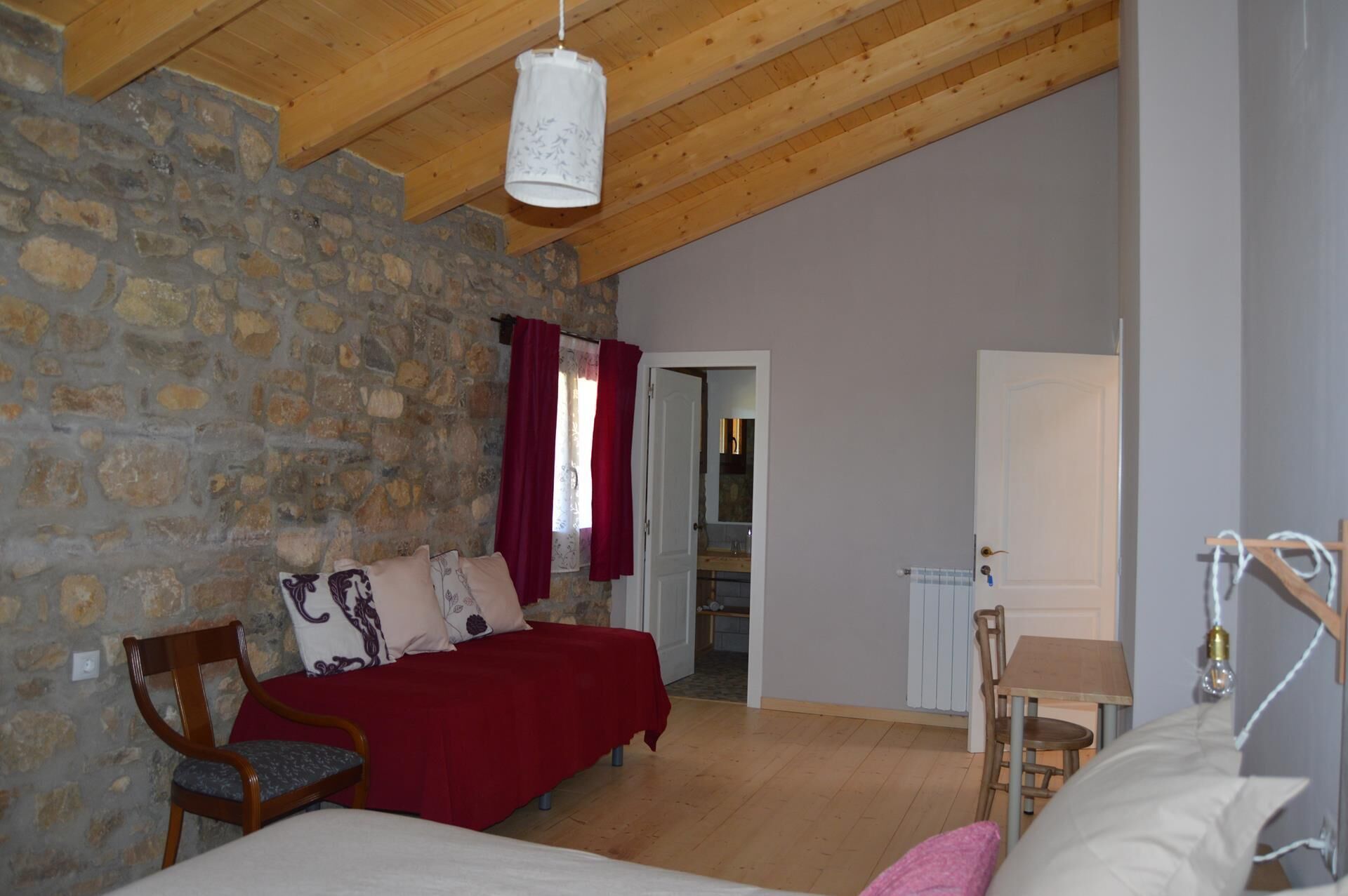 A bedroom with a stone wall, wooden ceiling beams, a bed with red bedding, a wooden chair, and a bedside table with a lamp.