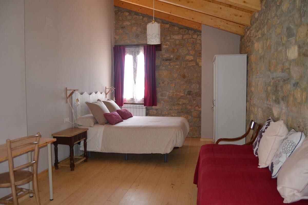 Quarto com parede de pedra, cama com cabeceira de madeira, cadeira de madeira, sofá vermelho e janela com cortinas.