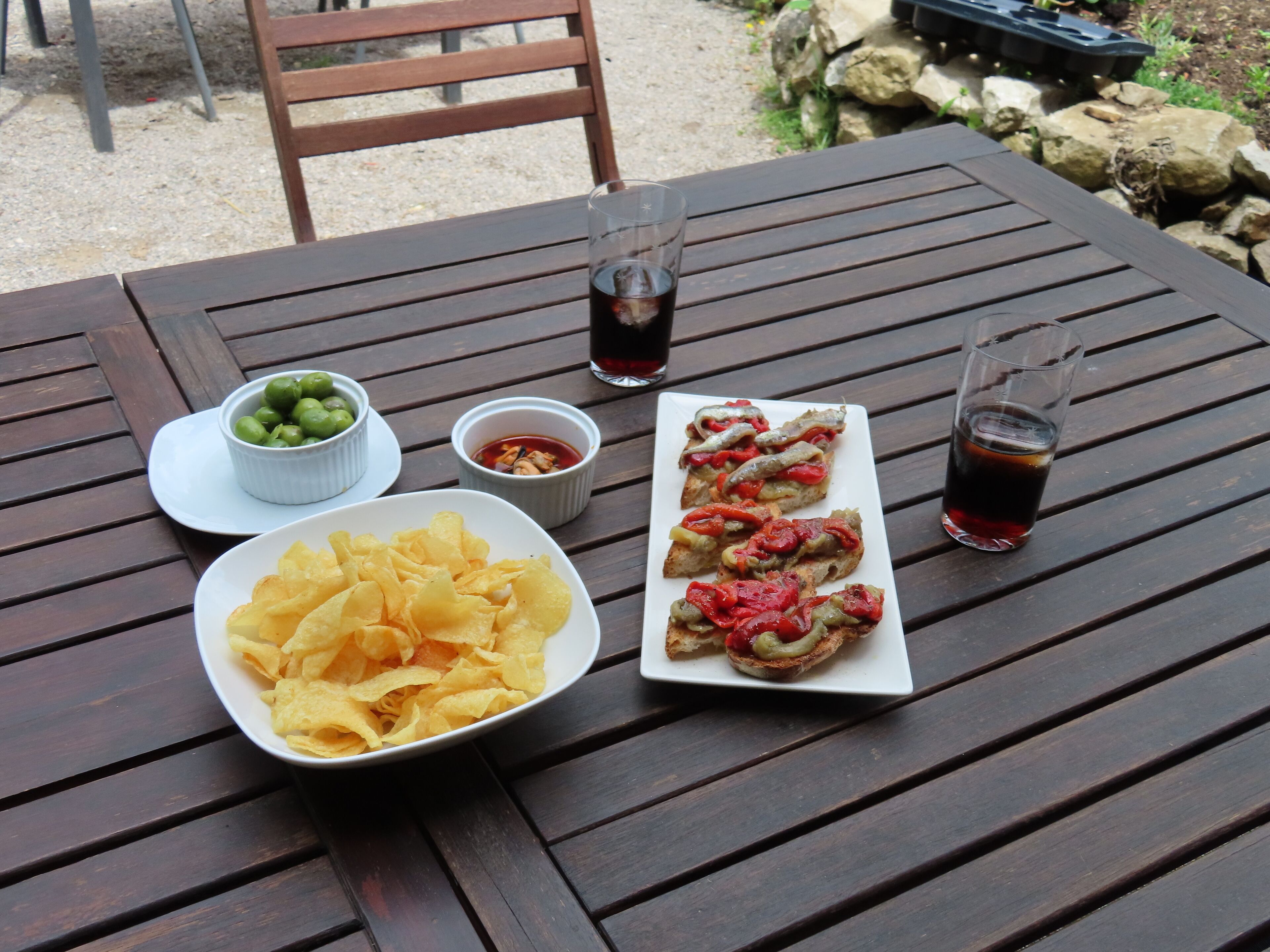 Una mesa de madera con un plato de papas fritas, un bol de aceitunas, un tazón pequeño de salsa y dos vasos de una bebida oscura.