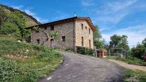 A stone building with a sloped roof, a paved path leading to it, and a fenced area with trees and shrubs. - Lo Cel De Tolo (Gavet de la Conca)