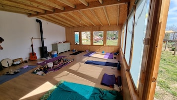 Amplia habitación con vigas de madera, ventanas grandes, colchonetas de yoga, una guitarra, una estufa y un armario.