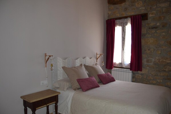 Un dormitorio con pared de piedra, cama con ropa de cama blanca y roja, y mesita de noche de madera.