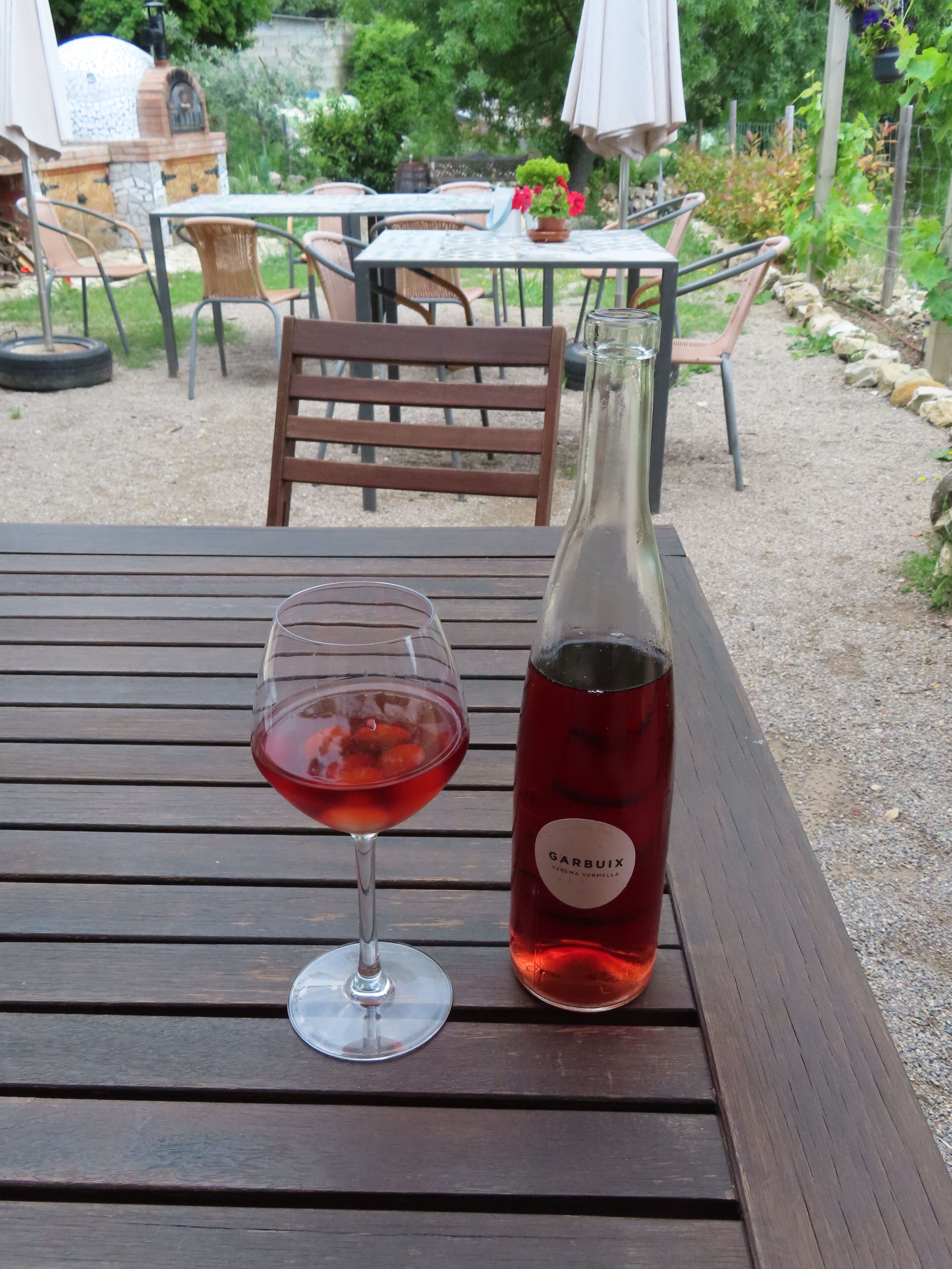 A bottle of red wine and a glass on a wooden table in an outdoor setting.