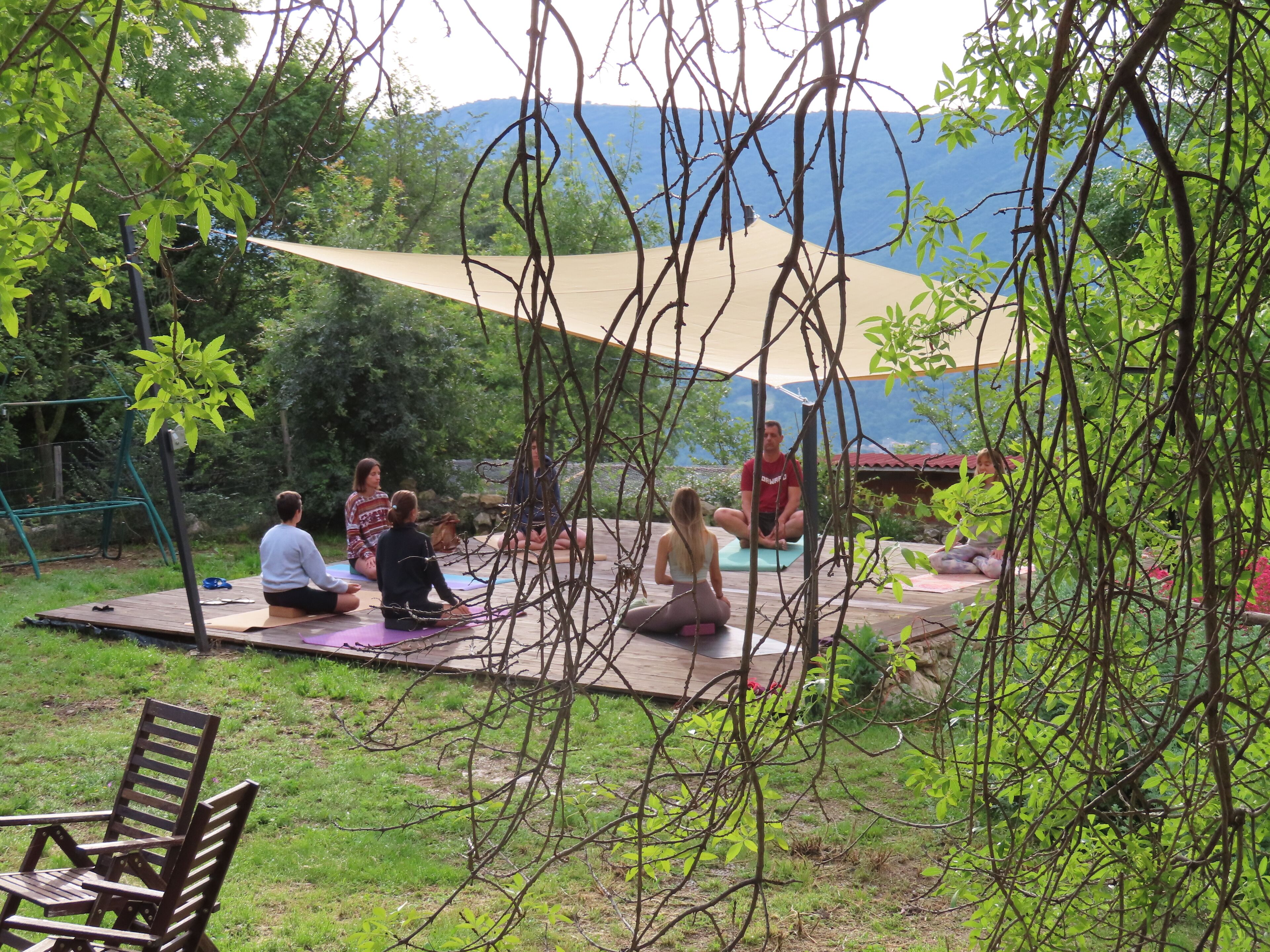 Un grupo de personas sentadas sobre esterillas al aire libre, quizás para una sesión de yoga o meditación, con una terraza de madera y un columpio al fondo.