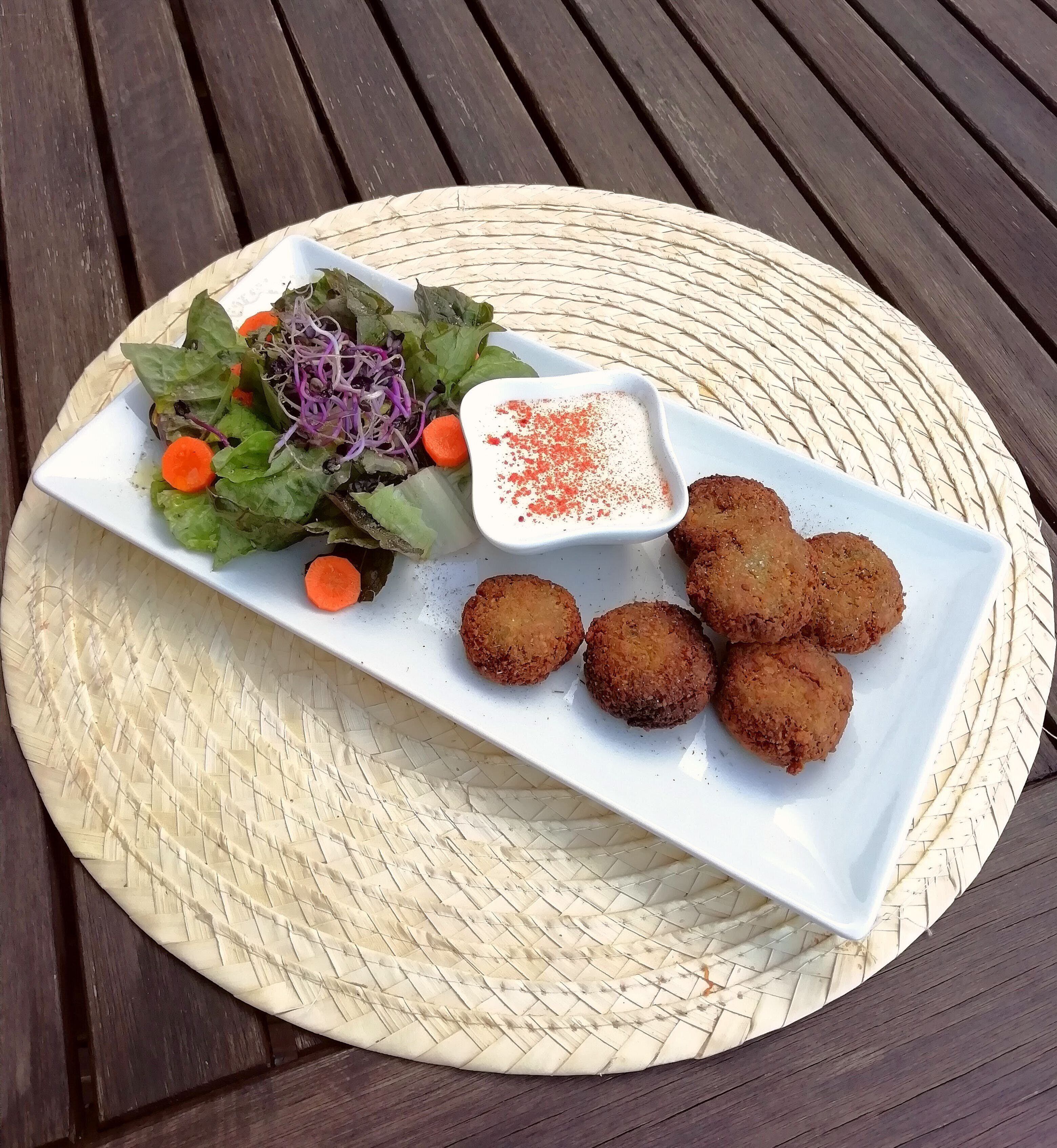 A white rectangular plate with a salad, a small bowl of sauce, and four fried items on a woven mat.