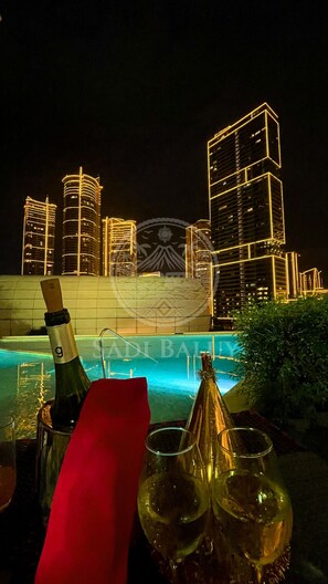 A nighttime view of a city skyline with illuminated buildings, a pool area with champagne and glasses, and a red napkin on a table. - Cozy Stay 3 min to Rockwell with City View, Pool, Parking (Mandaluyong City)