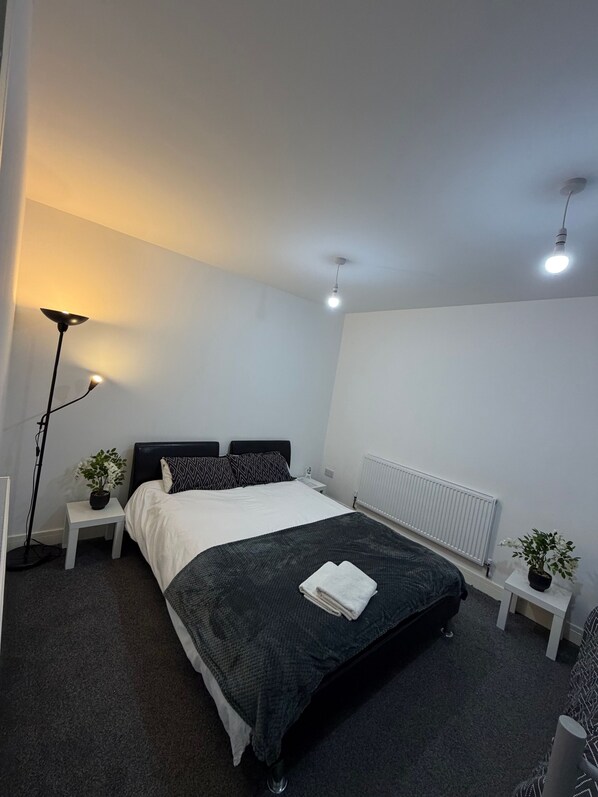 A bedroom with a bed, two bedside tables, a floor lamp, a radiator, and a potted plant.