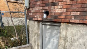 Un área de drenaje de concreto con una rejilla metálica, una puerta gris y un muro de ladrillo.