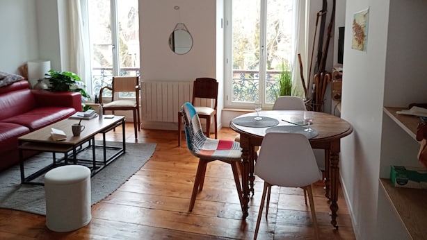 Ein Wohnzimmer mit einem roten Sofa, einem Couchtisch, einem Esstisch mit Stühlen, einem Heizkörper und einem Fenster mit Blick auf Grünflächen.