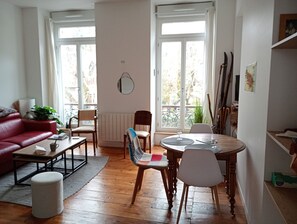 Un salon avec un canapé rouge, une table basse, une table à manger avec des chaises, un radiateur et une fenêtre donnant sur un espace verdoyant.