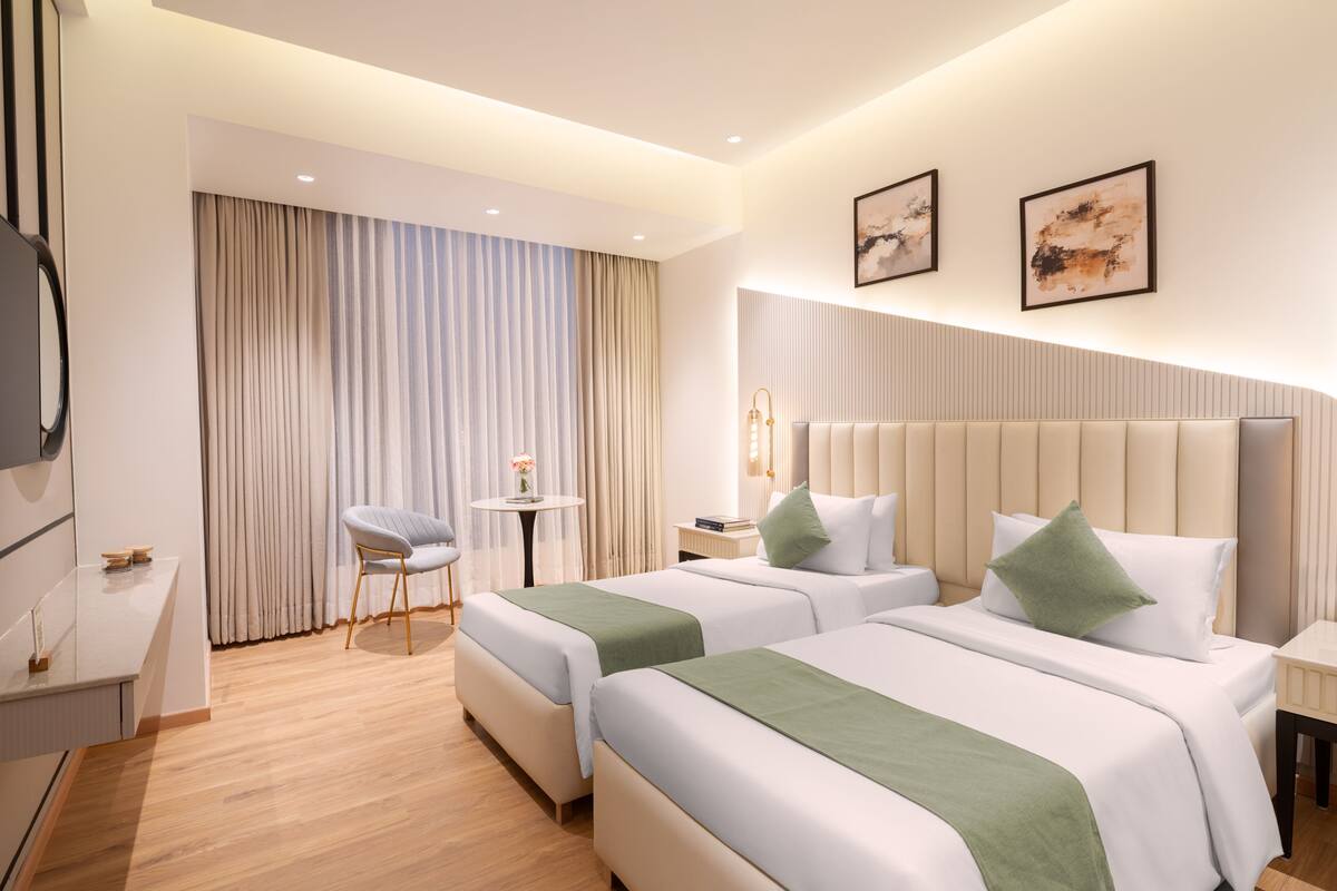 Une chambre d’hôtel avec deux lits, une petite table avec une chaise, une télévision et une fenêtre avec des rideaux.