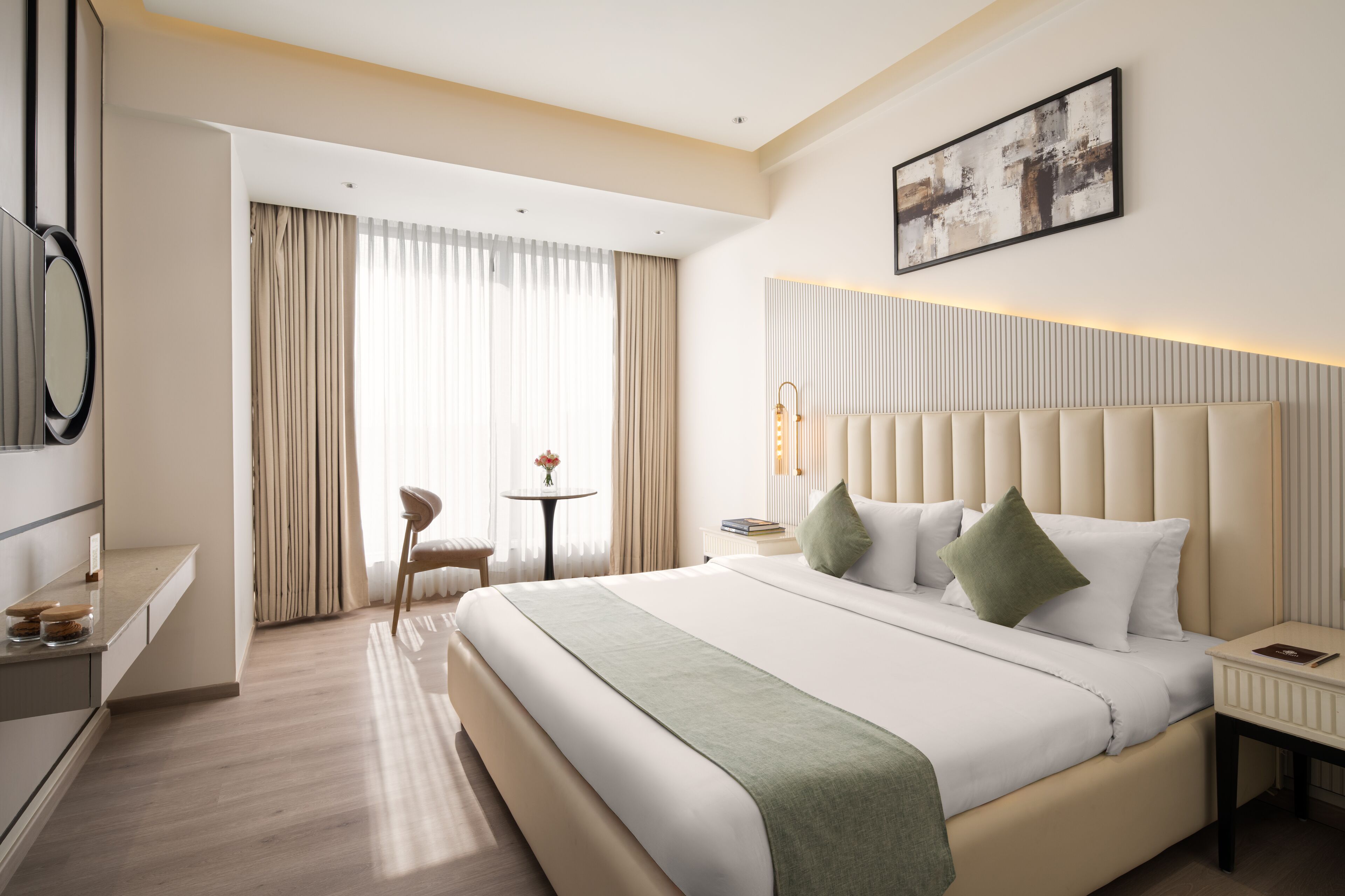 Una habitación de hotel moderna con una cama grande, una mesita de noche, una silla, una pequeña mesa con un jarrón y un ventanal con cortinas.