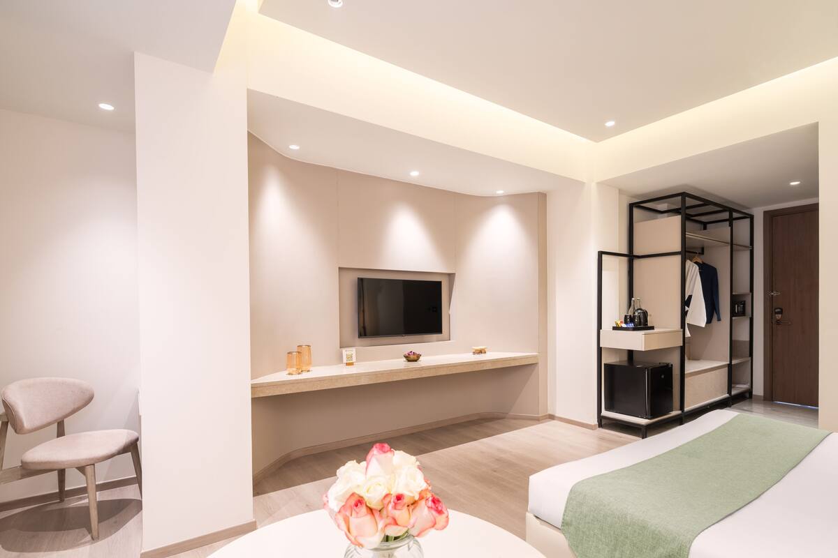 Une chambre d’hôtel moderne équipée d’un lit, d’un téléviseur fixé au mur, d’un coin salon avec un fauteuil et d’une coiffeuse avec un miroir et des étagères.