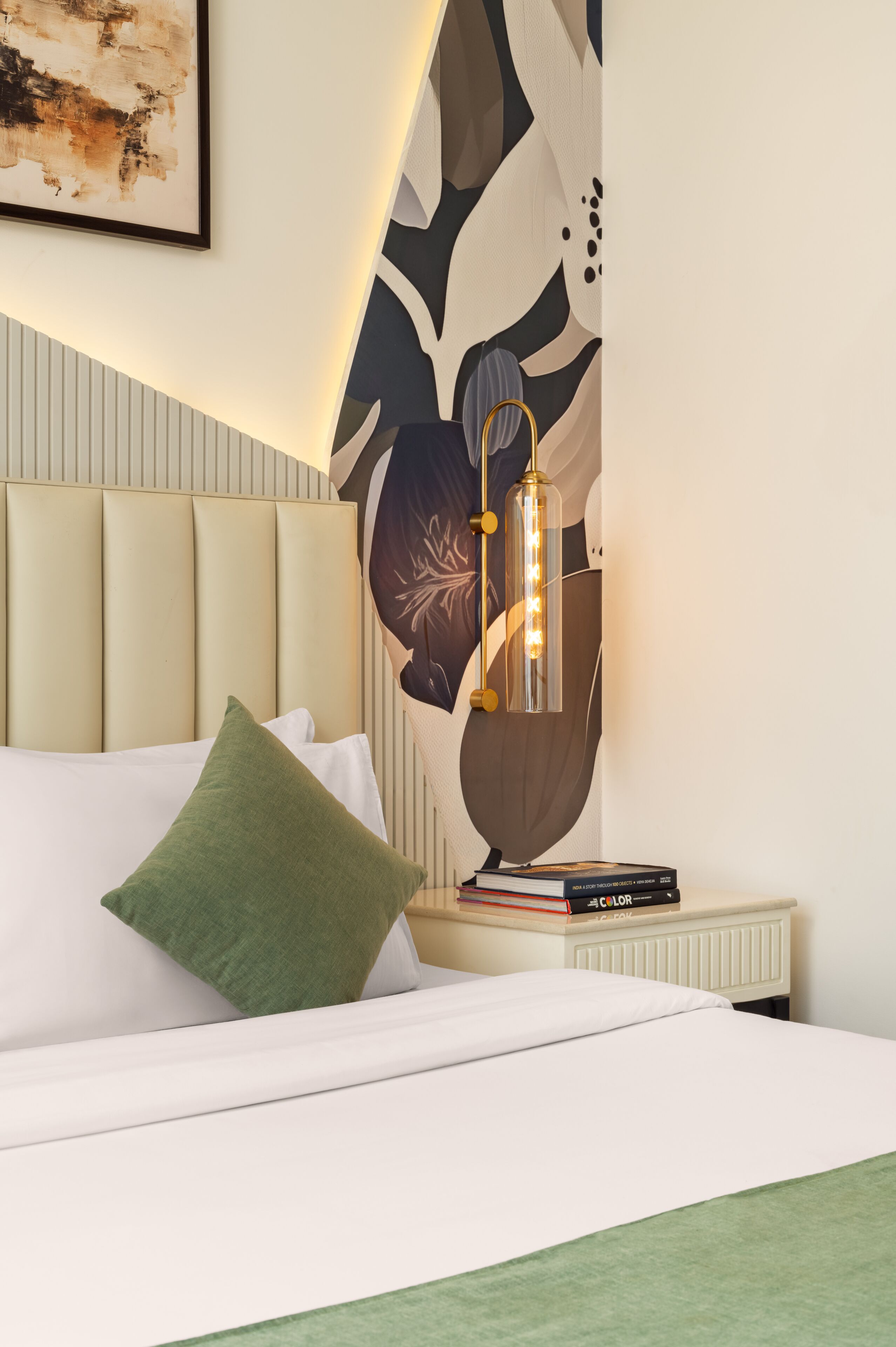 A neatly made bed with a green pillow, a bedside table with books, and a wall-mounted lamp.