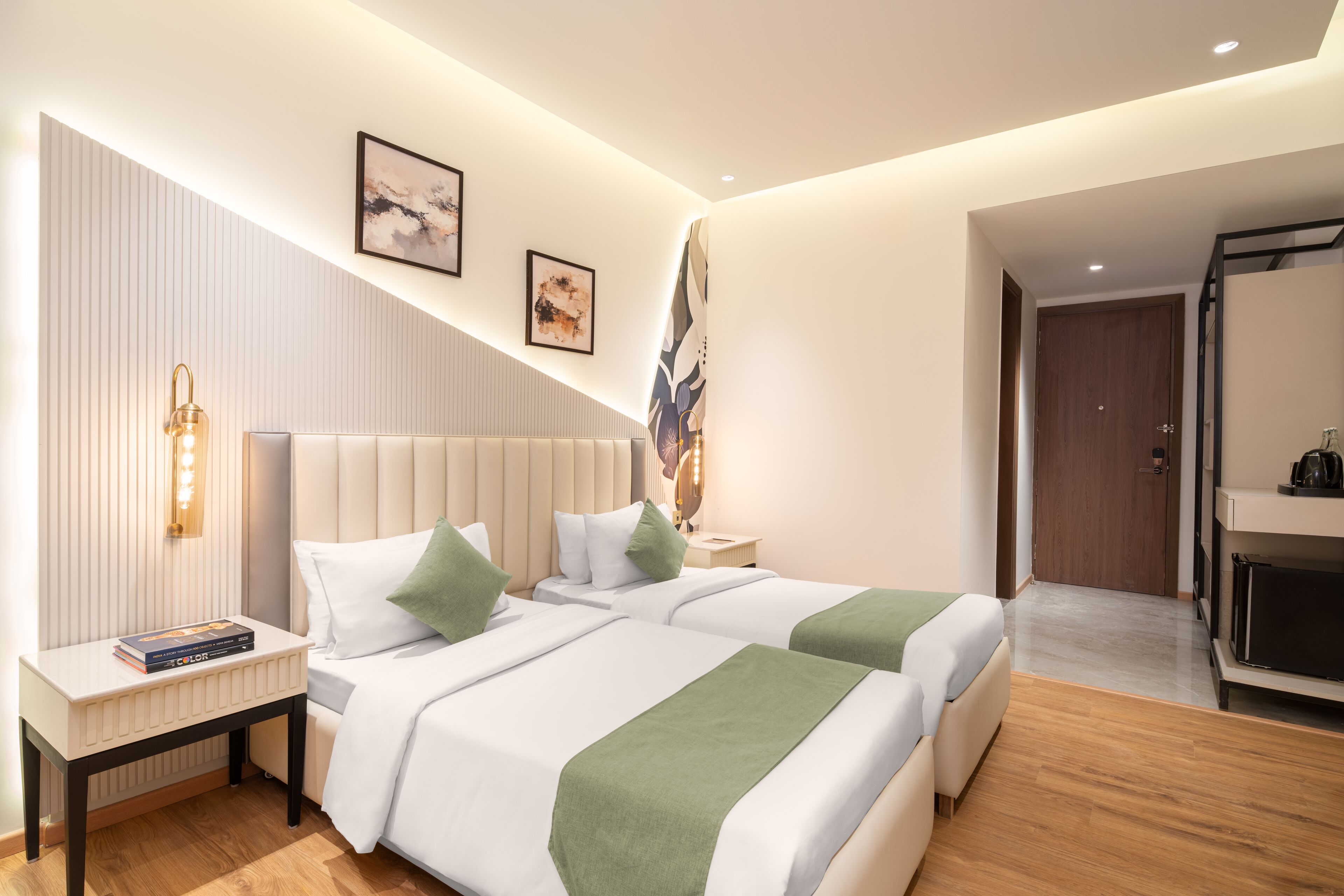A modern hotel room with two beds, a nightstand, a wall-mounted light, and a door leading to a bathroom.