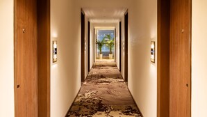 A hotel hallway with wooden doors, a patterned carpet, and a view of palm trees through the windows. - Foxtrail Sector 60- AM Hotel Kollection (Sohna)