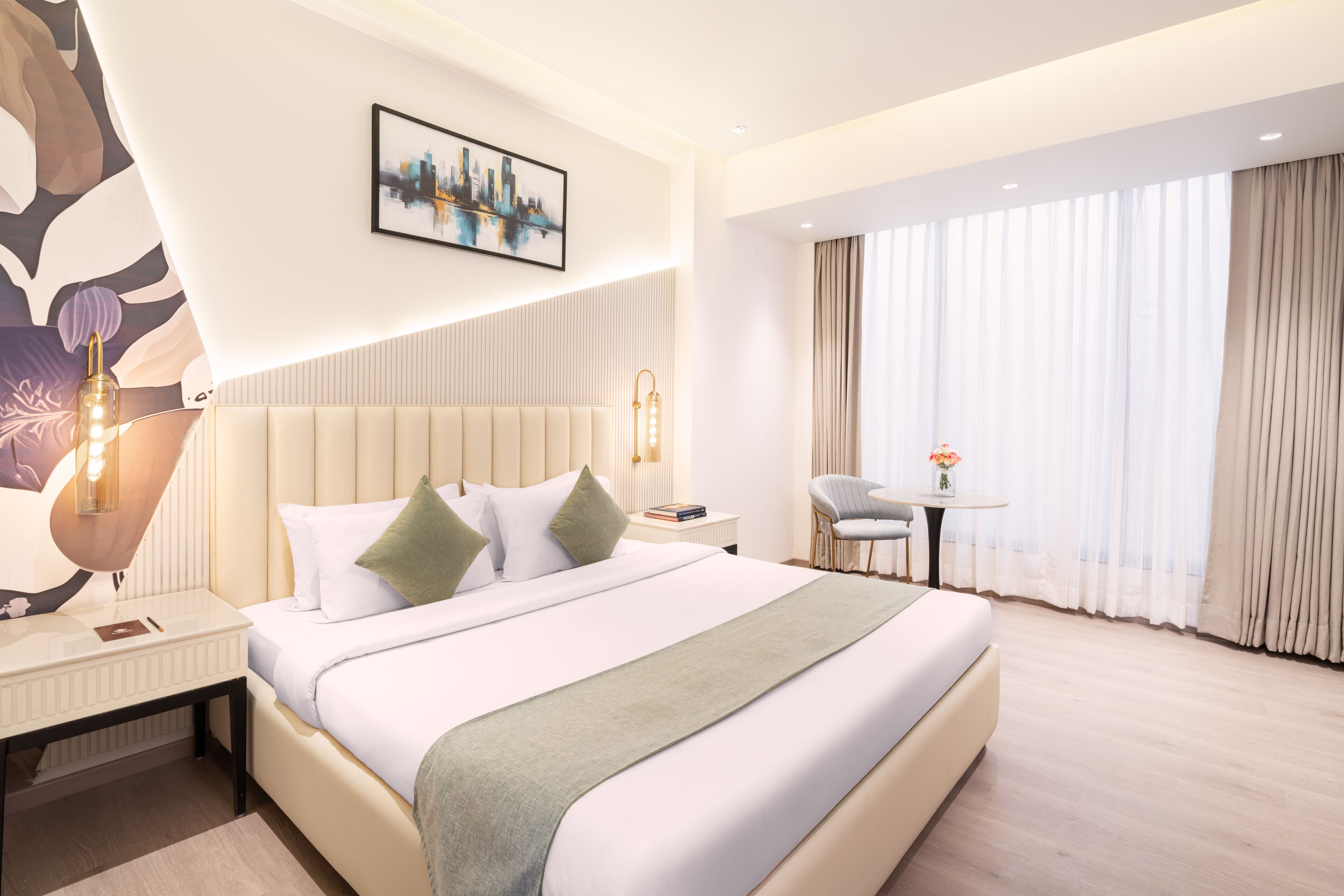 Quarto de hotel moderno com uma cama grande, uma mesinha de cabeceira, uma cadeira, uma pequena mesa com um vaso de flores e uma pintura de paisagem urbana na parede.