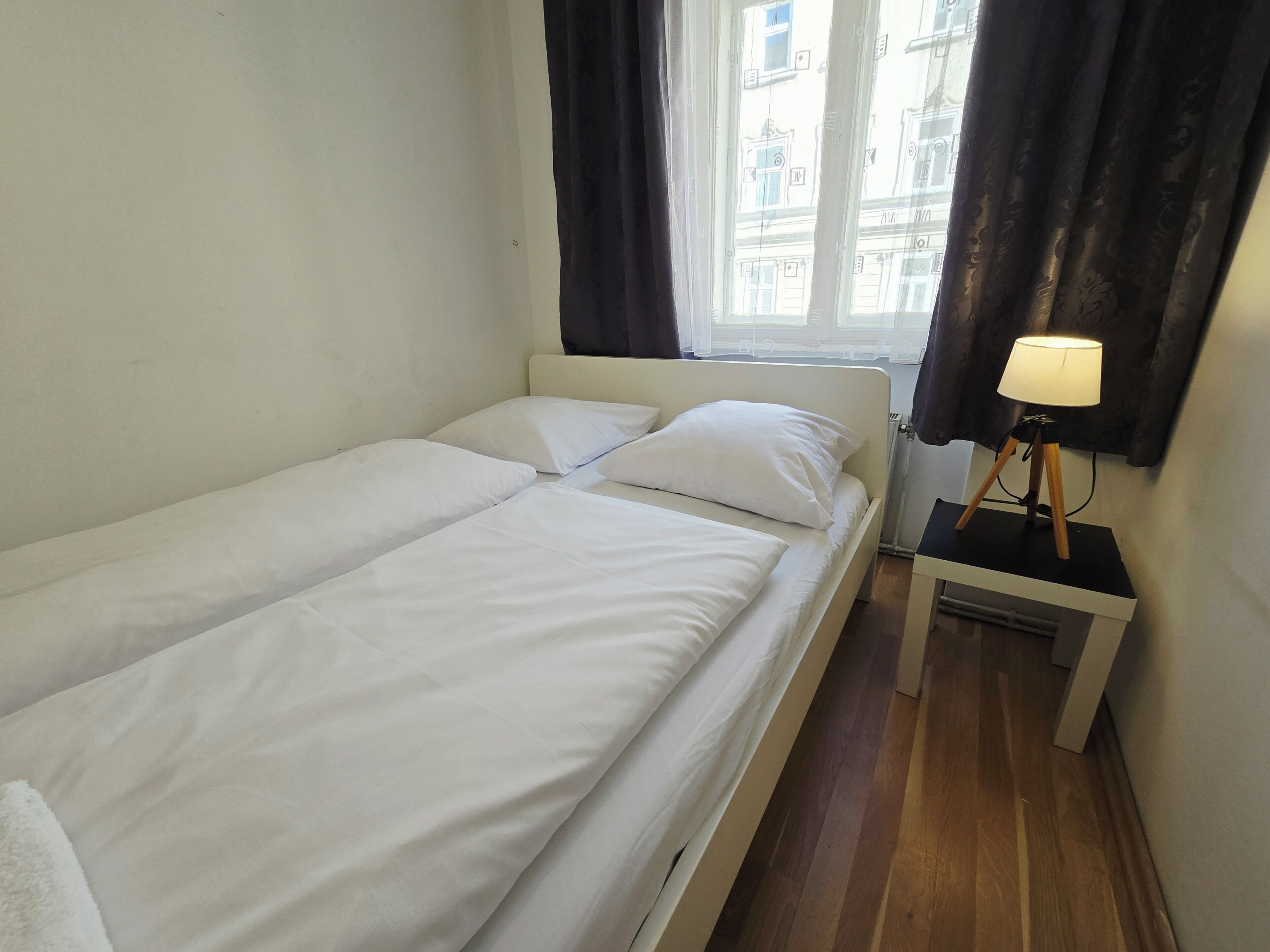 Una habitación pequeña y limpia con dos camas individuales, una mesita de noche con lámpara y una ventana con cortinas.