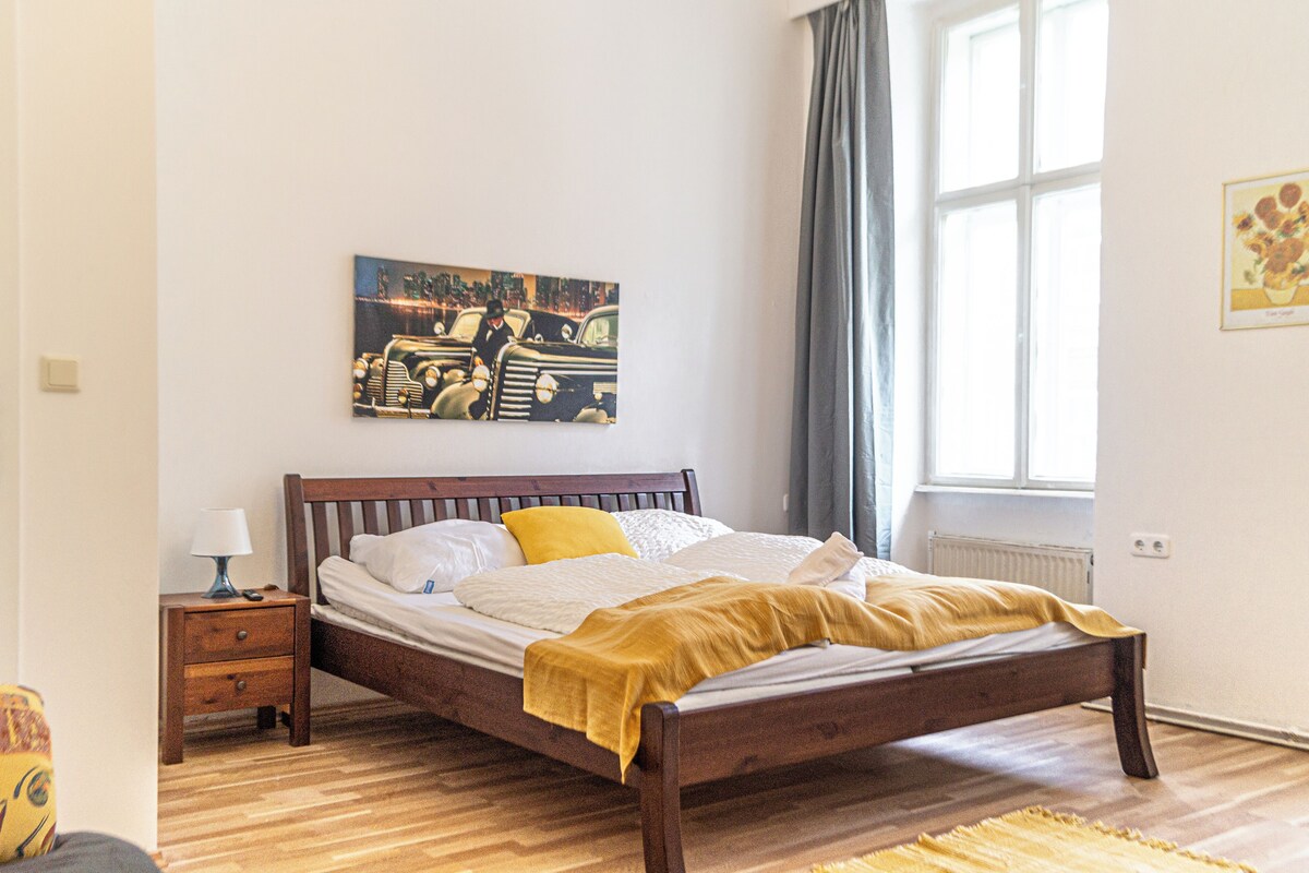 A bedroom with a wooden bed, a nightstand, a lamp, a painting on the wall, and a window with curtains.