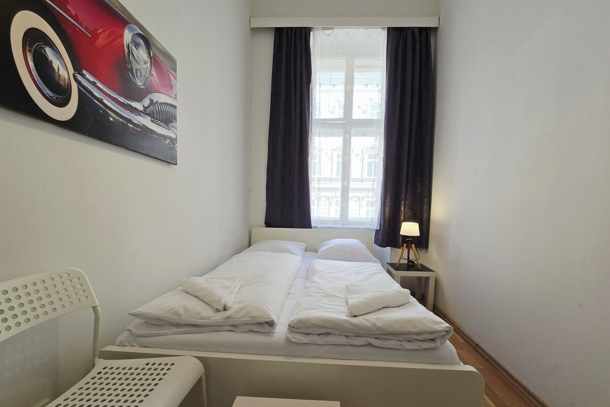A hotel room with a bed, a small table, a chair, a window with curtains, and a wall-mounted lamp.