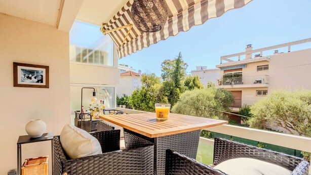 Un balcone con mobili in vimini, un tavolo con un bicchiere di succo d'arancia e vista su edifici e alberi circostanti.
