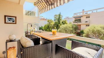 A balcony with wicker furniture, a table with a glass of orange juice, and a view of neighboring buildings and trees.